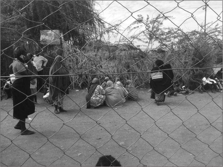 View through a chicken wire fence. A dry dirt path runs between the fence and a line of trees and scrub. Around 25 black people are queuing, mostly sitting on the ground, some standing. One woman is on a mobile phone. In the center of the picture, seven woman are squatting in a circle, all wearing brightly colored chadors/headscarves.
