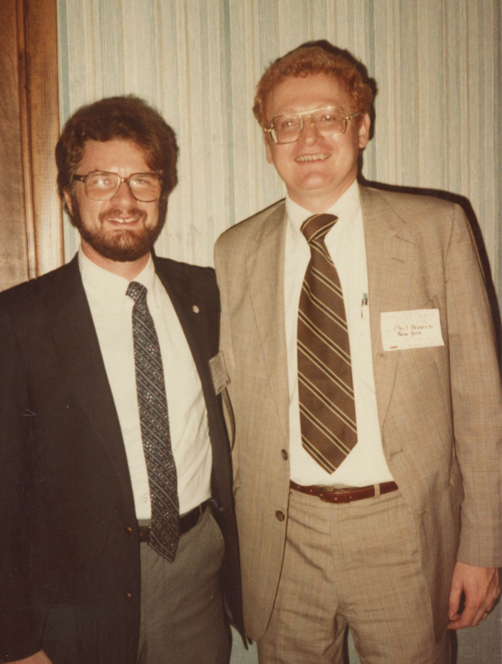 Two young white men in suits. The man on the left has dark hair and a mustache and beard. He's wearing a dark suit jacket with a narrow striped tie and has glasses. The other man has thick red haird and is wearing a light brown suit with a brown striped tie and a name tag reading Phil Bravin.
