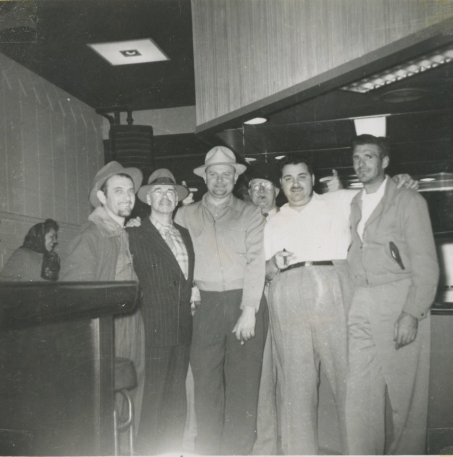 A group of six white men are standing at a bar. Three are wearing fedoras. Behind them is a woman with a scarf tied around her head.