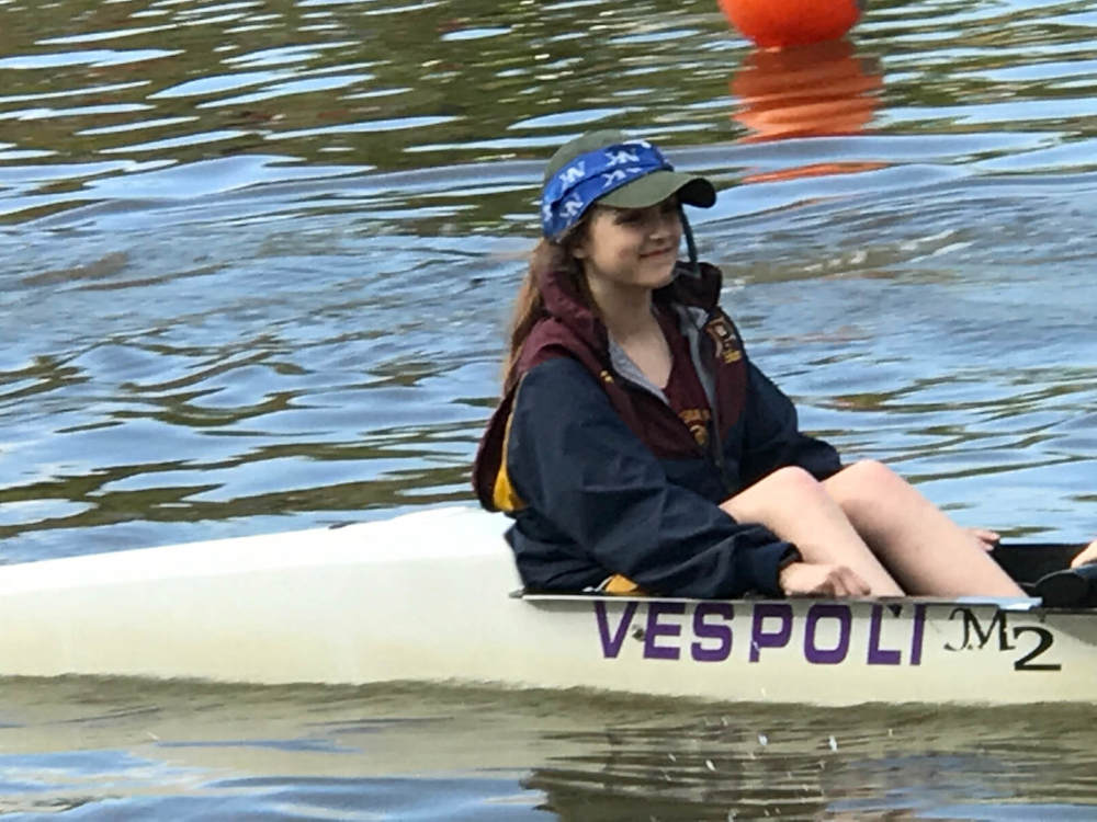 Young white girl with a baseball cap and a windbreaker sits in the coxswain seat of a rowing shell on the water of the Erie Canal. The words VESPOLI M2 are on the side of the boat.