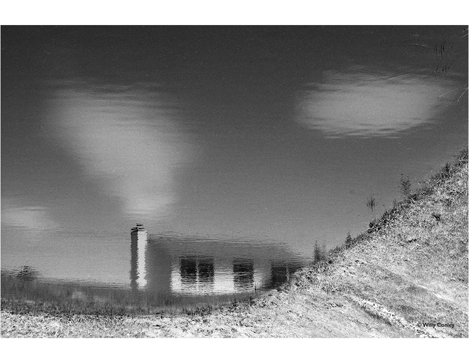 black-and-white horizontal photo of a large water reflection taken along the edge of a storm water management pond. The main subject in the reflection is a ranch house with a chimney that looks like smoke is coming out of it when in an otherwise clear sky are clouds floating by. The edge of the pond gives the reflected house image the appearance of being on a prairie with a hill.