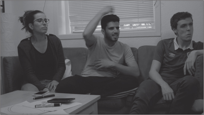 Three young people in casual clothes are sitting on a sofa, with a coffee table in front of them with pens, markers, paper, a wallet and a phone strewn on it. On the left, a young white woman with glasses, her long brown hair tied back, is watching someone off camera. In the middle, a young Middle Eastern man with dark hair and a goatee is signing, his right hand raised above his head, blurred with movement. His left elbow is bent, and his left hand sits at his chest level. On the right, a young white man with short brown hair looks at someone off-camera.