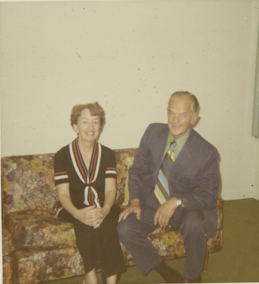 A middle-aged white man and woman sit on a brown and yellow floral patterned couch. The woman has her legs crossed and her hands are clasped around her knee. She has on a black dress with red and white accents around the neckline and the cuffs of its short sleeves. She has short brown wavy hair. The man is in a gray suit with a green shirt and a wide, striped tie. He has receding white hair and a tan.
