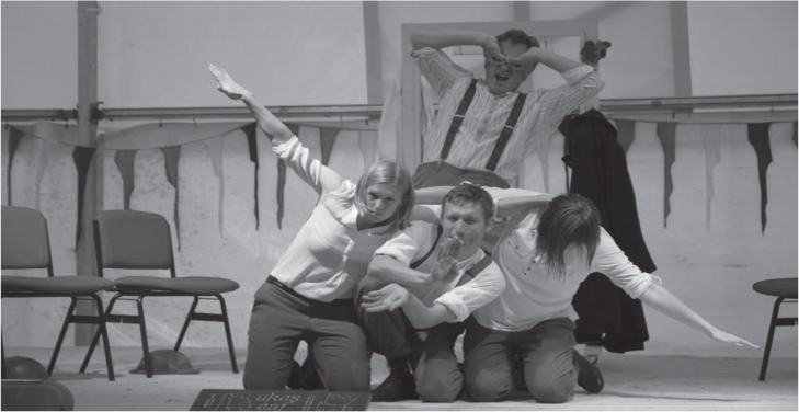 Three students sit on the floor. The young man in the middle acts like the nose of the plane. The students on either side of him each have one arm outstretched, mimicking the wings of a plane. Sitting behind them, the fourth students acts like the pilot.
