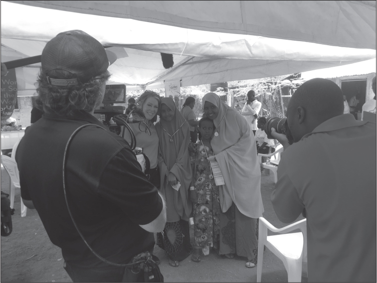 Interior of a tent. The camera is behind a white male camera operator in a baseball cap, and a black bald man in a T-shirt with a long-lens camera, who are filming/photographing a group of four people: a white smiling woman in a bright top, a young smiling black girl in a bright chador/head scarf, and a younger black girl in a long patterned dress, being hugged by a smiling black woman in a bright chador/head scarf.