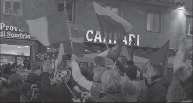 A group of predominantly or exclusively white people in winter clothing flying standing in the street (backs to camera) flying various flags.