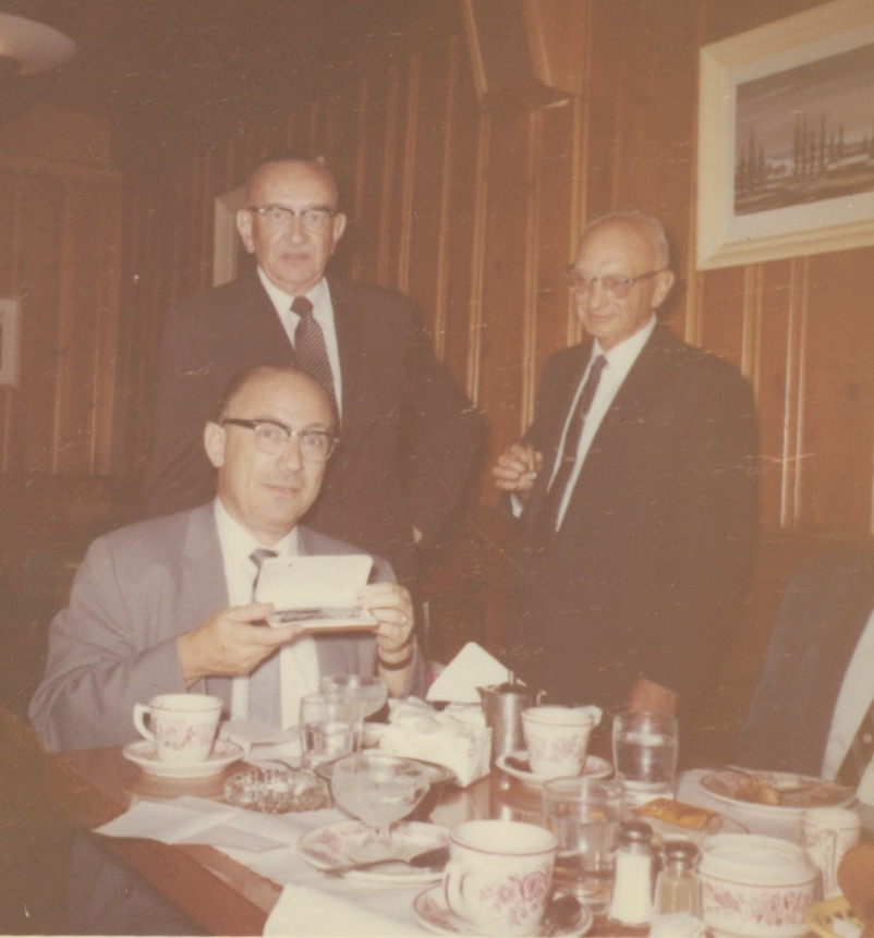 An older white man wearing a light gray suit with horn-rimmed glasses and a receding hairline is seated at a table. He's holding up a gift box with a watch inside it. Behind him, standing, are two older white men both wearing glasses and in suits. On the table are mugs and waterglasses as if a meal had just been finished.