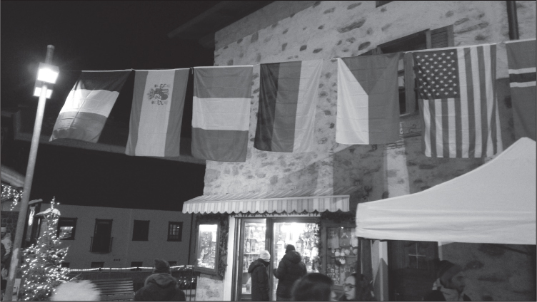 Nighttime exterior of an Italian building. Various flags (French, Spanish, Italian, German, American and Icelandic?) are hanging like bunting across the road, while people mill beneath,
