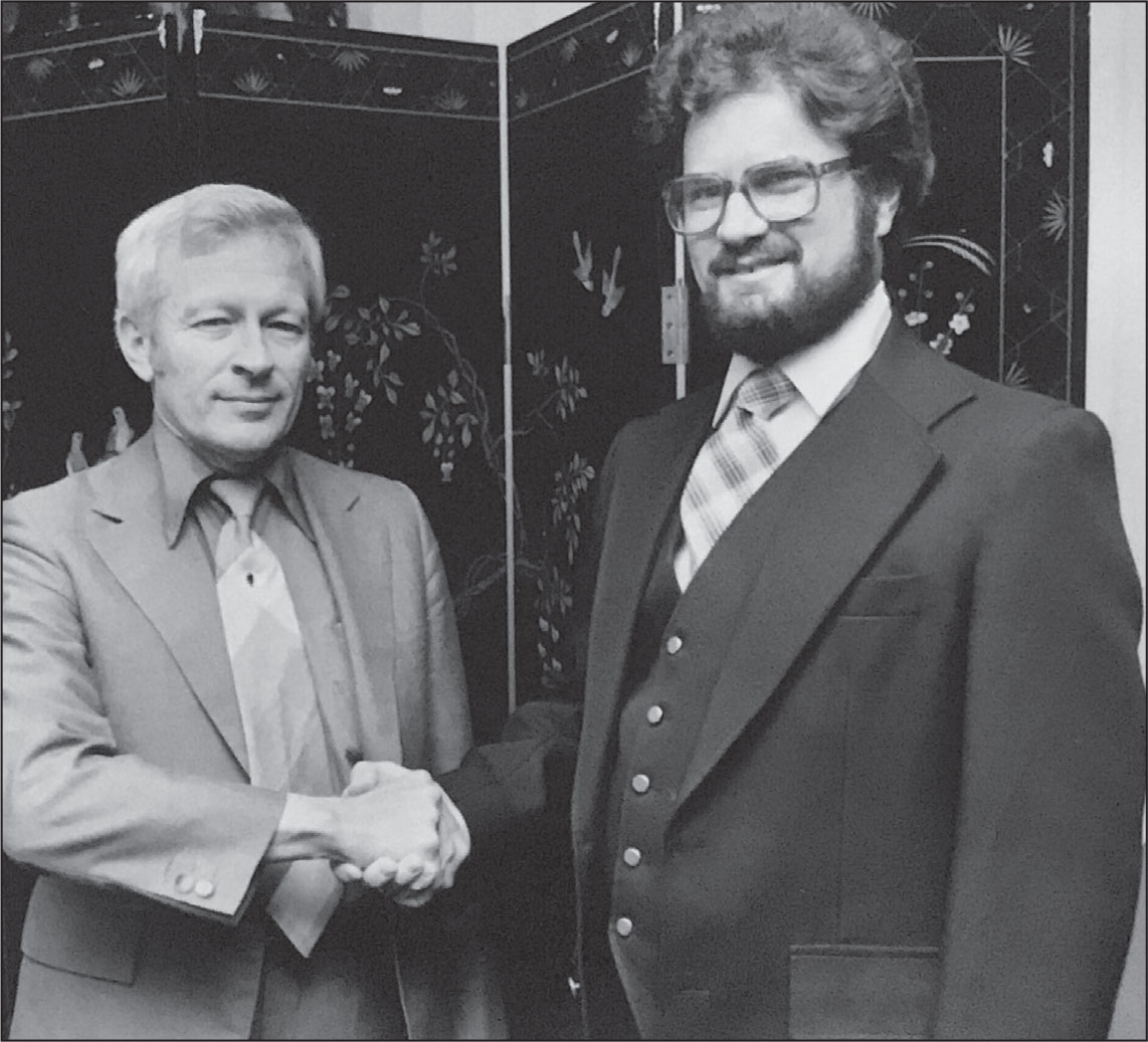 An older white man in a light-colored suit shakes the hands of a young white man in a dark suite. They's standing in front of a dark screen painted with flowers and birds.