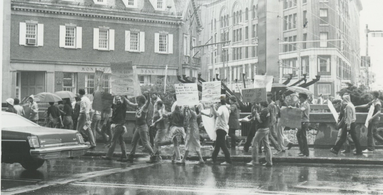 A black-and-white photo of a large group of people holding up signs walking toward a building in thei rain.
