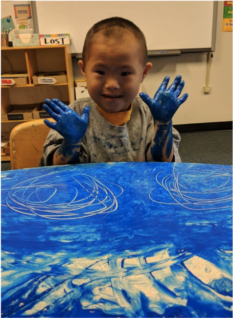 A young boy wearing a paint-splattered smock sits in front of a table covered in swirls of blue paint. He holds both hands up--both are covered in blue paint.
