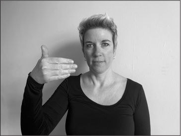 Annelies, a white woman with short blonde hair, signs FLAG (right hand in front of right shoulder, palm facing away from the camera, fingers extended but closed, thumb up).