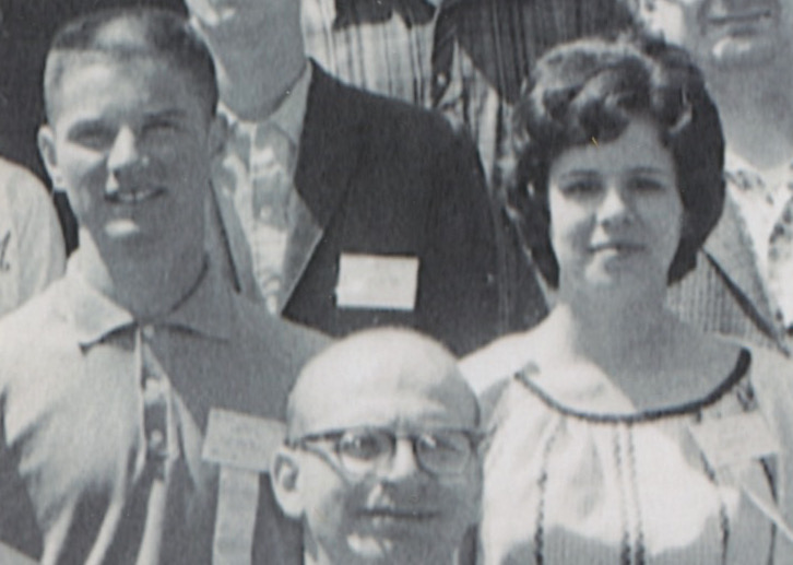A white young man with a crew cut stands in a crowd of people. He is wearing a buttoned up polo shirt and has a name tag on his chest. Next to him is a young white woman with thick dark hair styled in a bouffant. She also wears a name tag.