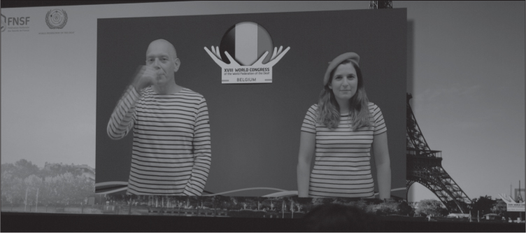 A screen above the stage at a congress, showing a white man and woman wearing blue and white striped tops; the woman has a red beret. The man is signing BELGIUM (right hand in a fist in front of mouth). Between them is a logo showing two hands holding up a circle containing the Belgian flag, with text reading XVIII WORLD CONGRESS of the World Federation of the Deaf BELGIUM.