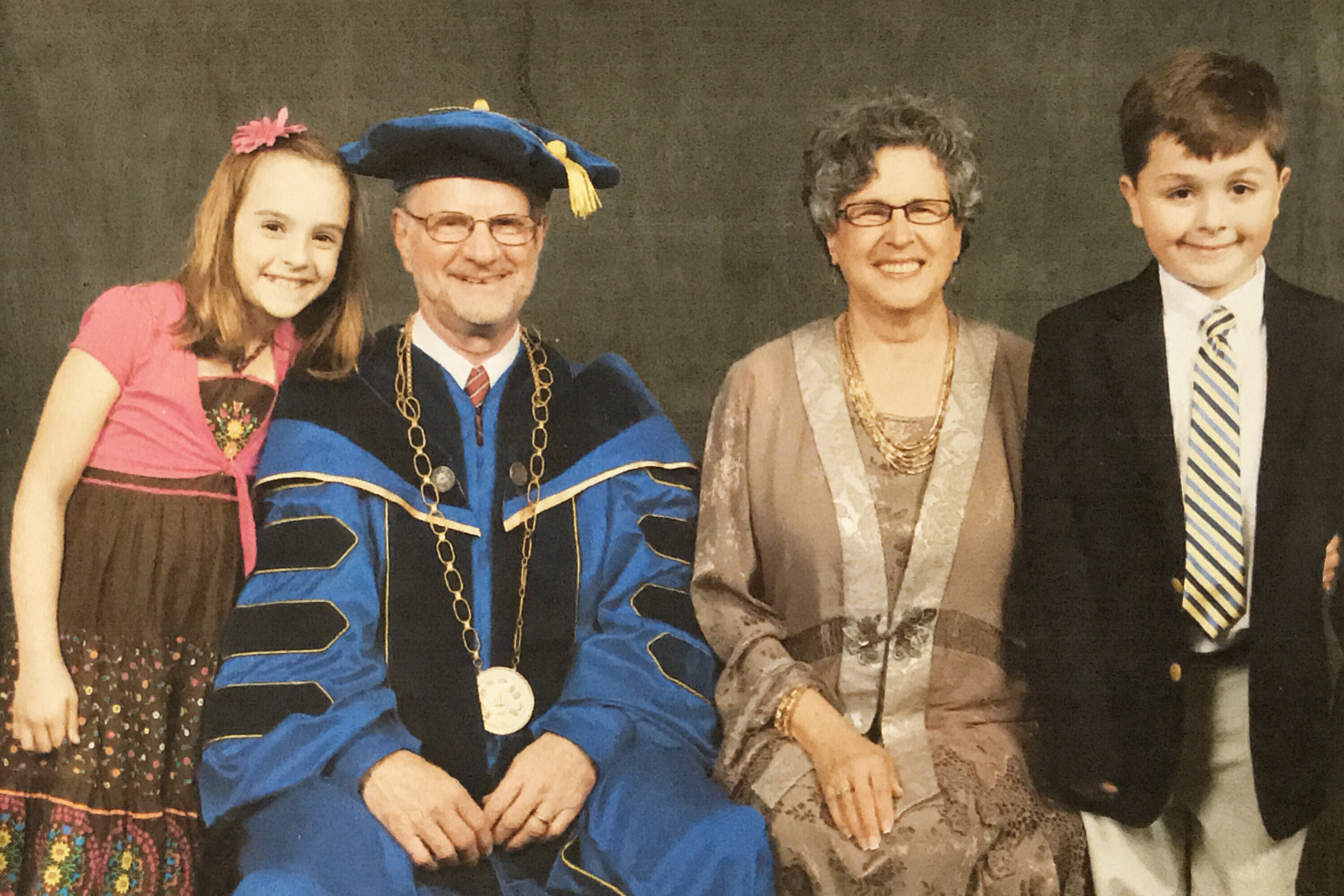 A young white girl in a pink and brown dress with a pink ribbon in her long hair leans on the shoulder of her grandfather, a white older man in a blue doctoral robe with a heavy pendant around his neck. Next to him sits an older white woman with wavy gray hair and glasses, wearing a taupe and light brown dress. A young white boy in a dark navy blazer and striped tie stands next to her.