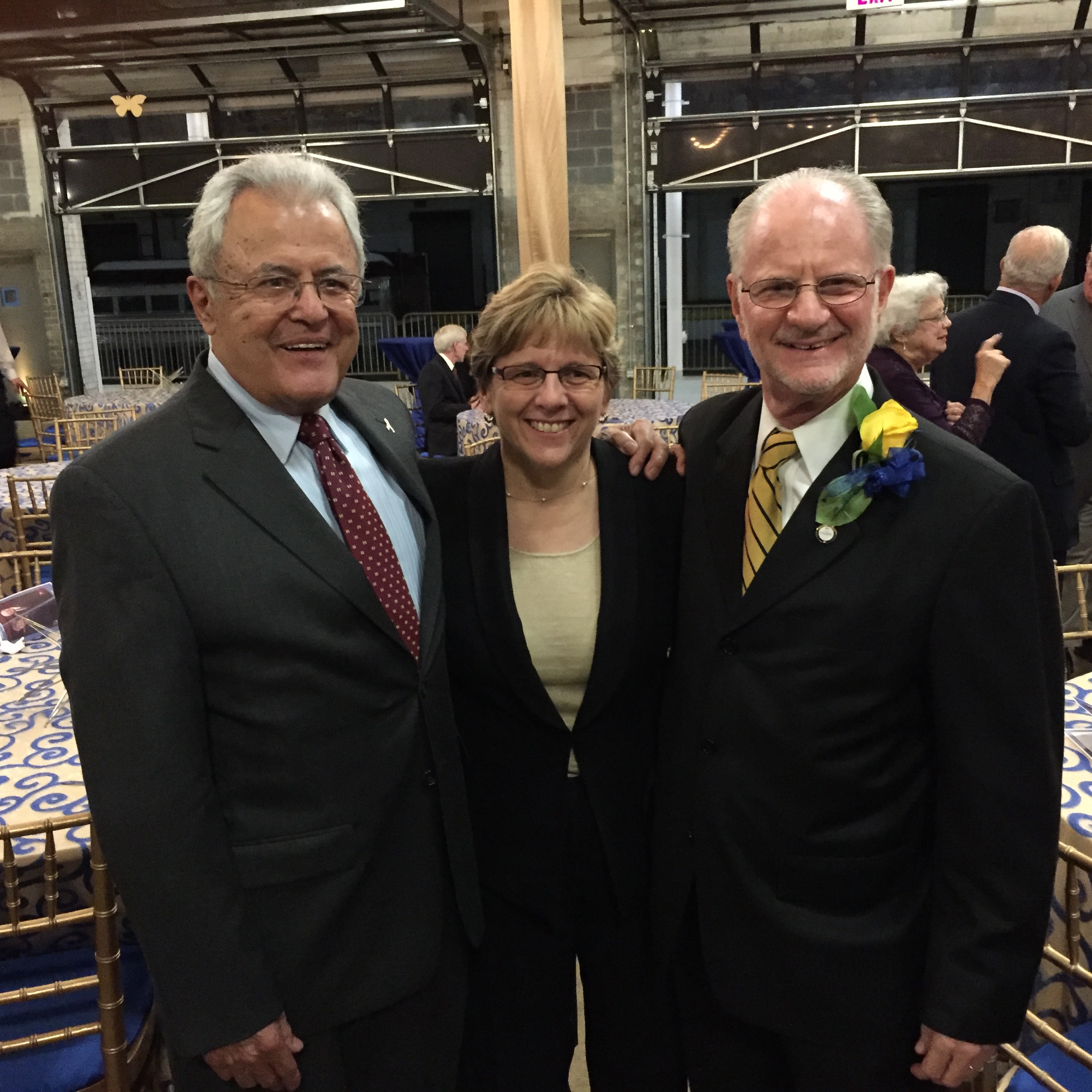 An older Latino man in a dark navy suit and maroon spotted tie has his arm around a short middle-aged white woman with short blond hair and glasses. She's wearing a dark jacket over a light green top. Standing next to her is an older white man with white hair and beard in a dark suit with a yellow and blue striped tie. A yellow rose is pinned to his lapel. Behind them tables are set up for a party.