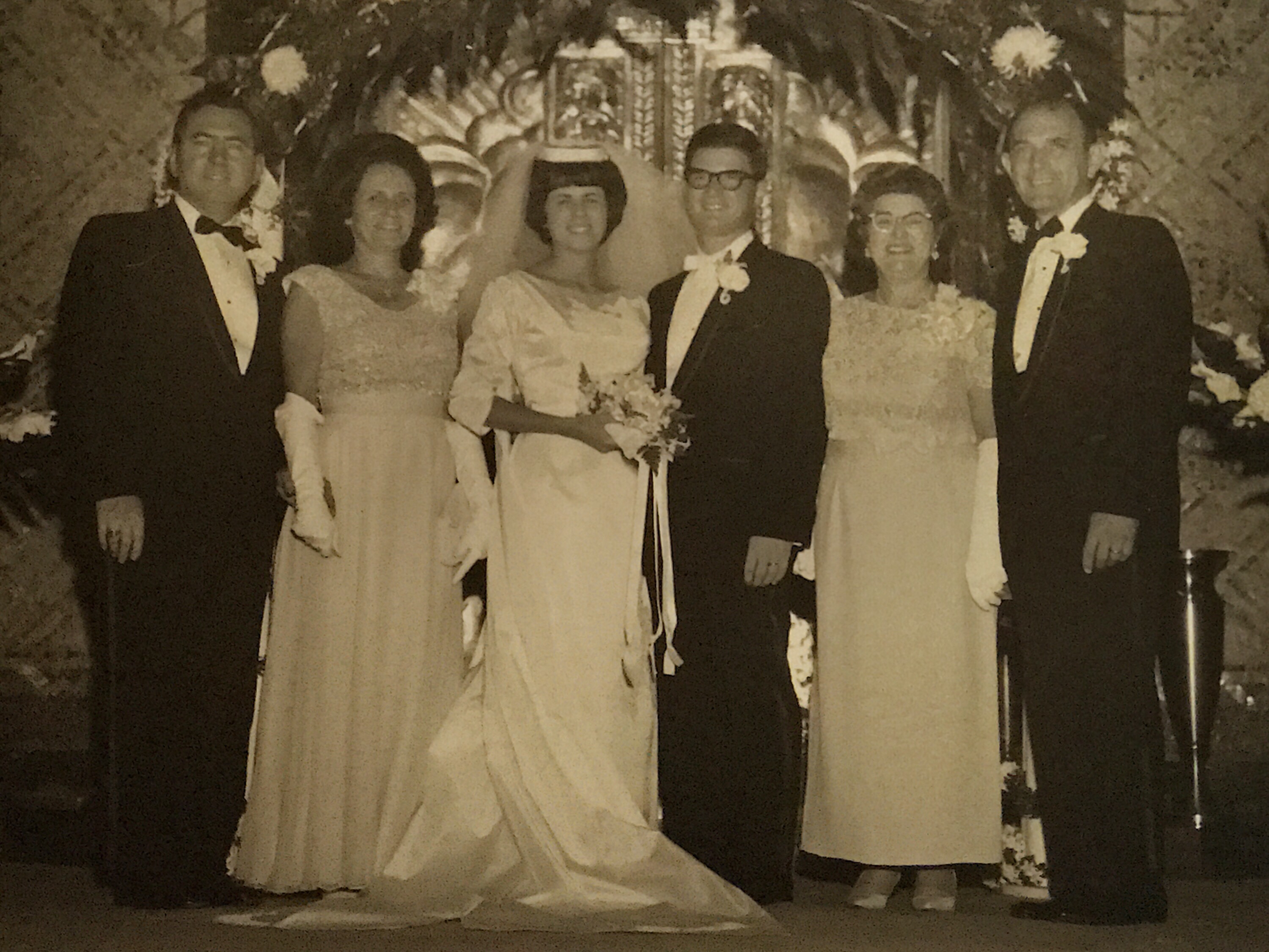 A formal photo of the bride and groom with their parents. The bride and groom are in the center of the photo. The men are all wearing tuxedos and the women are all in long dresses.