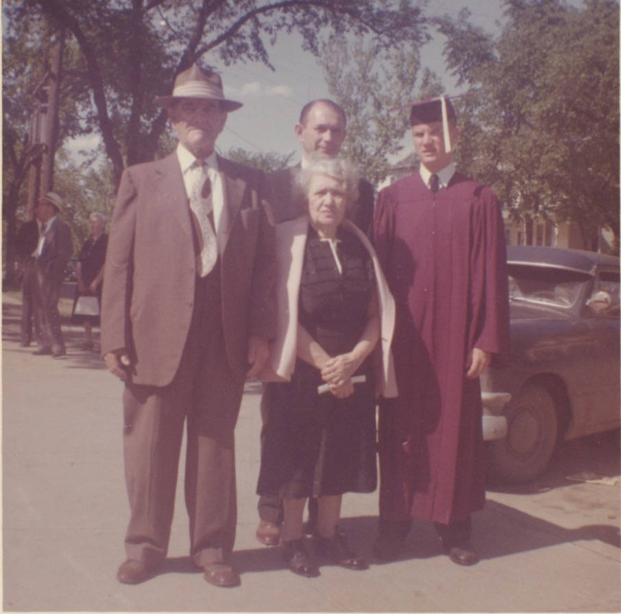 Four white people stand outside in front of a car. The first man is tall and wears a baggy brown suit and a fedora. Next to him is an older white woman with gray hair, wearing a black dress with a white shawl. Behind her is a middle-aged white man with a receding hairline. Next to him is a tall teenage boy wearing a maroon robe and a black mortar board with a tassle.