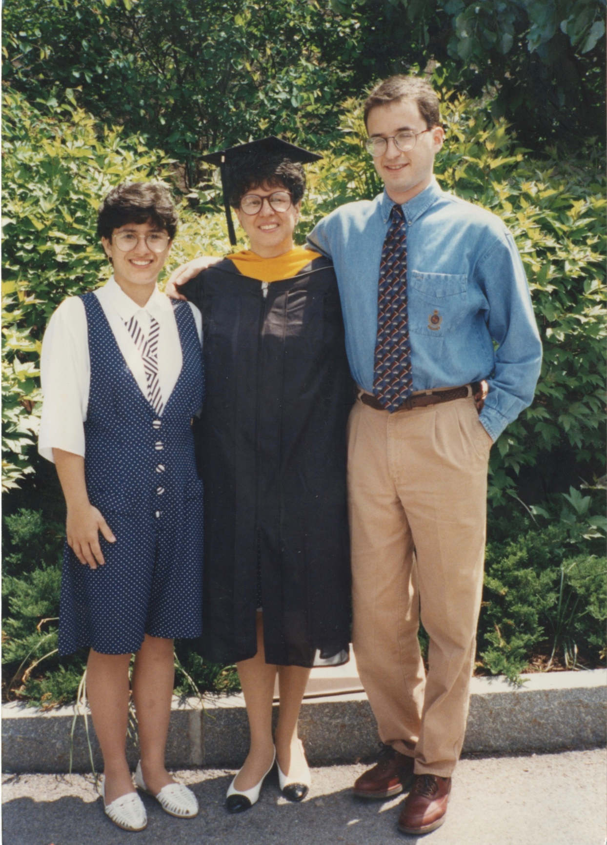 A middle-aged woman wearing a black robe and mortar board stands in front of some green bushes with her arm around a young girl with dark hair wearing a jumper with a white blouse underneath. On her other side is a tall, slim young man with light brown hair in a blue shirt with a patterened tie and khakis.