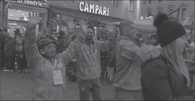 Three white men in green hoodies walking down a commercial street waving with both arms at spectators behind crash barriers.