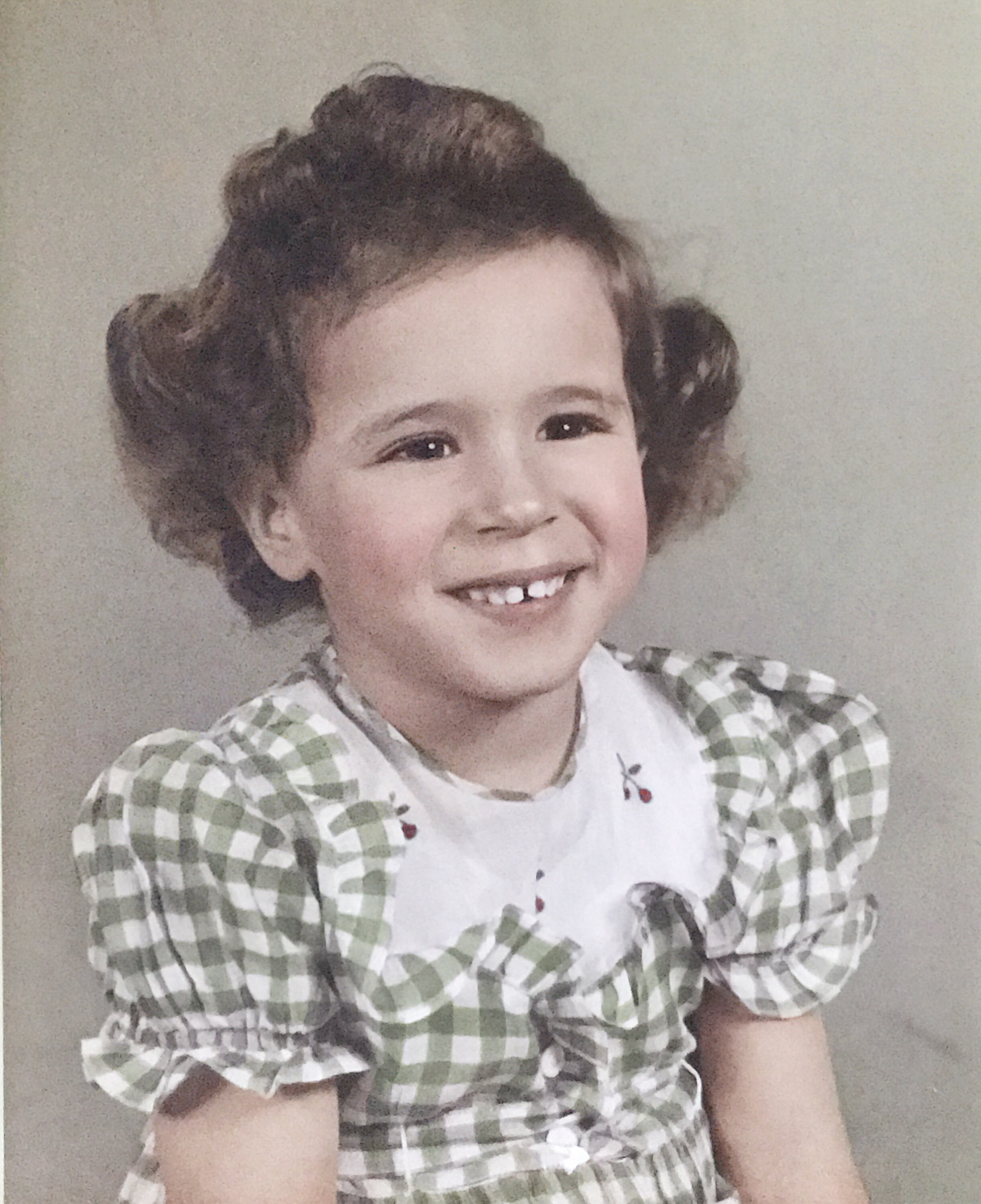 A little white girl with a gap-toothed smile looks off to the right. She has wavy dark hair and is wearing a green checkered dress with puffy sleeves. The collar of her dress is white and has red flowers embroidered on it.