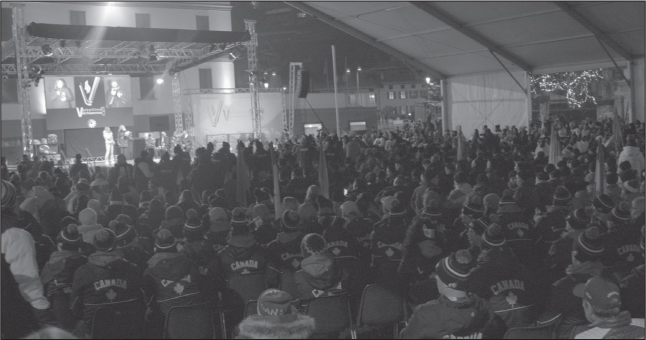 A large temporary shelter in a commercial street. Two people are standing and five or six sitting on a stage at the far end, which has a lighting rig and a screen. The shelter is packed with seated people in winter clothing facing the stage. Some are holding flags. A large proportion are wearing blue and red ski jackets with CANADA on the back. 