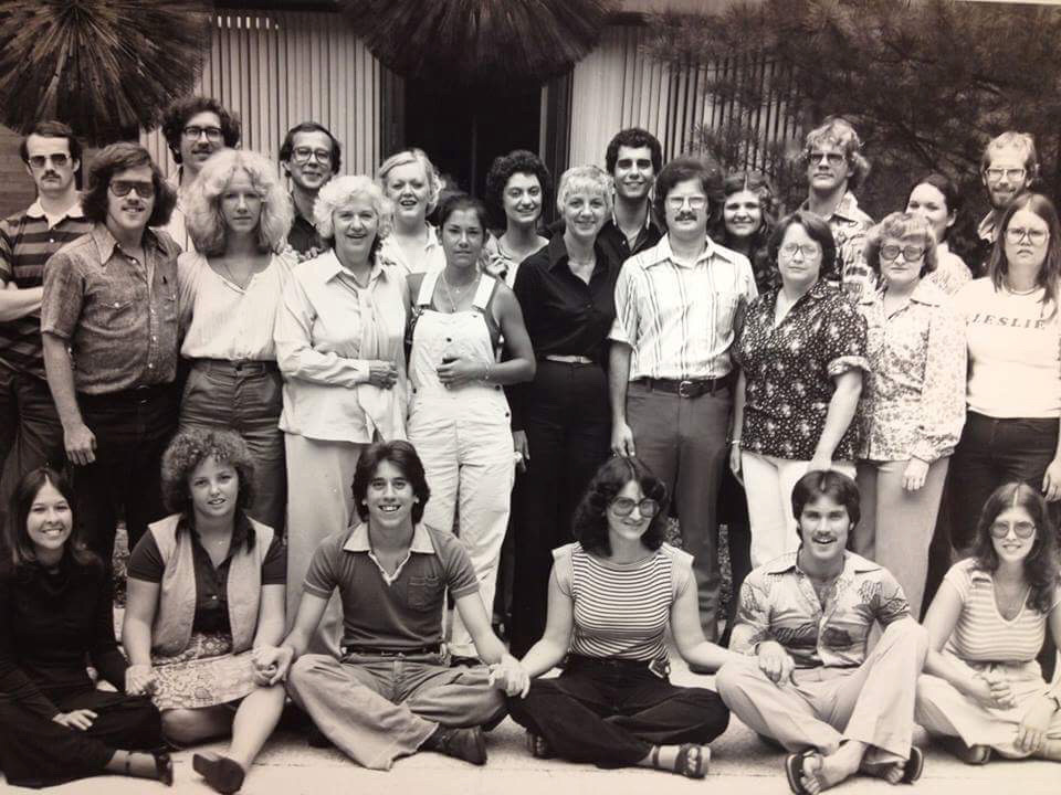 A large group of mostly young white people sitting and standing. The people in the front who are sitting on the floor are holding hands.