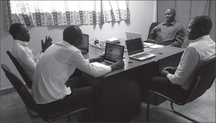 (currently 5.15) Interior of an office room with light green walls, beige floor, and African print curtains. Four black men in shirts and trousers sit around a table with laptops, signing.