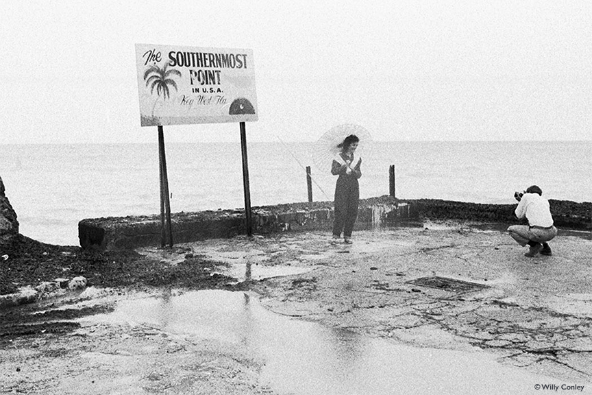 A black-and-white photo of a woman standing on a concrete pier holding a parasol. A man crouches a few feet away with a camera, taking a picture of her. Behind her is the ocean and a sign that reads 