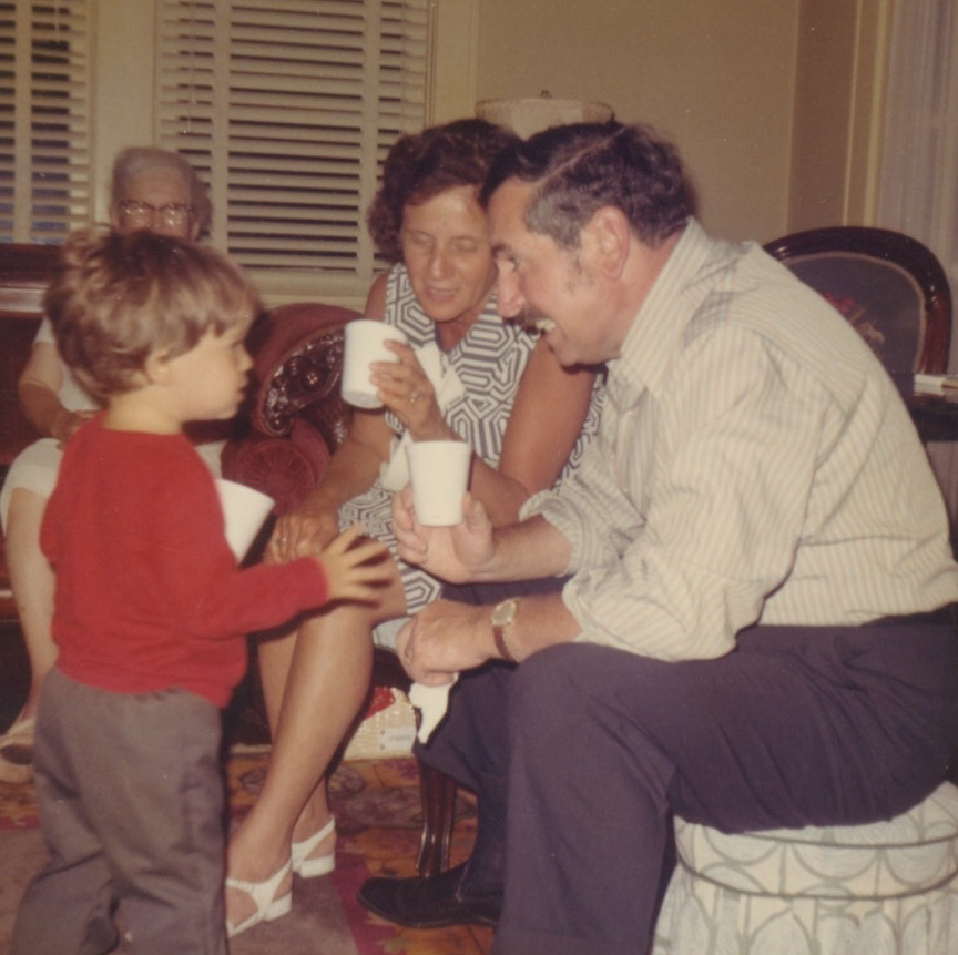 A young white boy in a red shirt and long pants holding a styrofoam cup stands in front of an older white man who is sittin on an ottoman. Next to him a middle-aged white woman leans in to join the conversation. In the background sits an elderly woman with white hair and glasses.