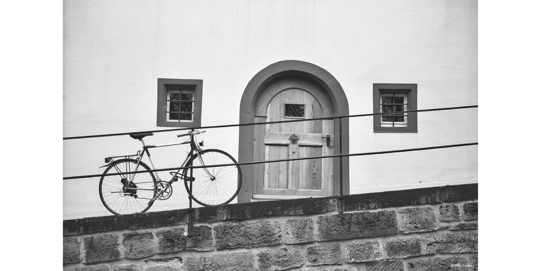 black-and-white horizontal photo of a 21-speed hybrid bike chained to a metal railing that goes along the top of an inclined rock wall. Behind the bike is a medieval-looking wood door with an arched top flanked by two small windows on each side. The windows of iron security bars over them.