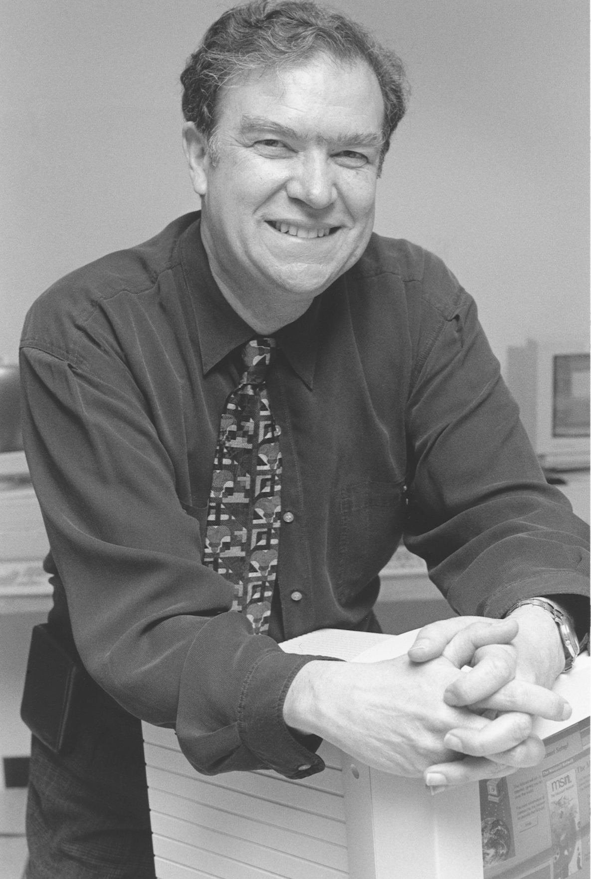 Black-and-white photo of a middle-aged white man in a dark button-up shirt with a patterned tie. He's leaning on a computer monitor with both hands clasped in front of him.