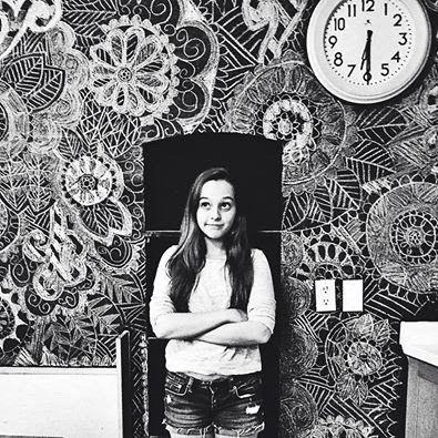 A young white girl with long dark hair stands in front of a wall covered in intricate patters of flowers and mandalas.