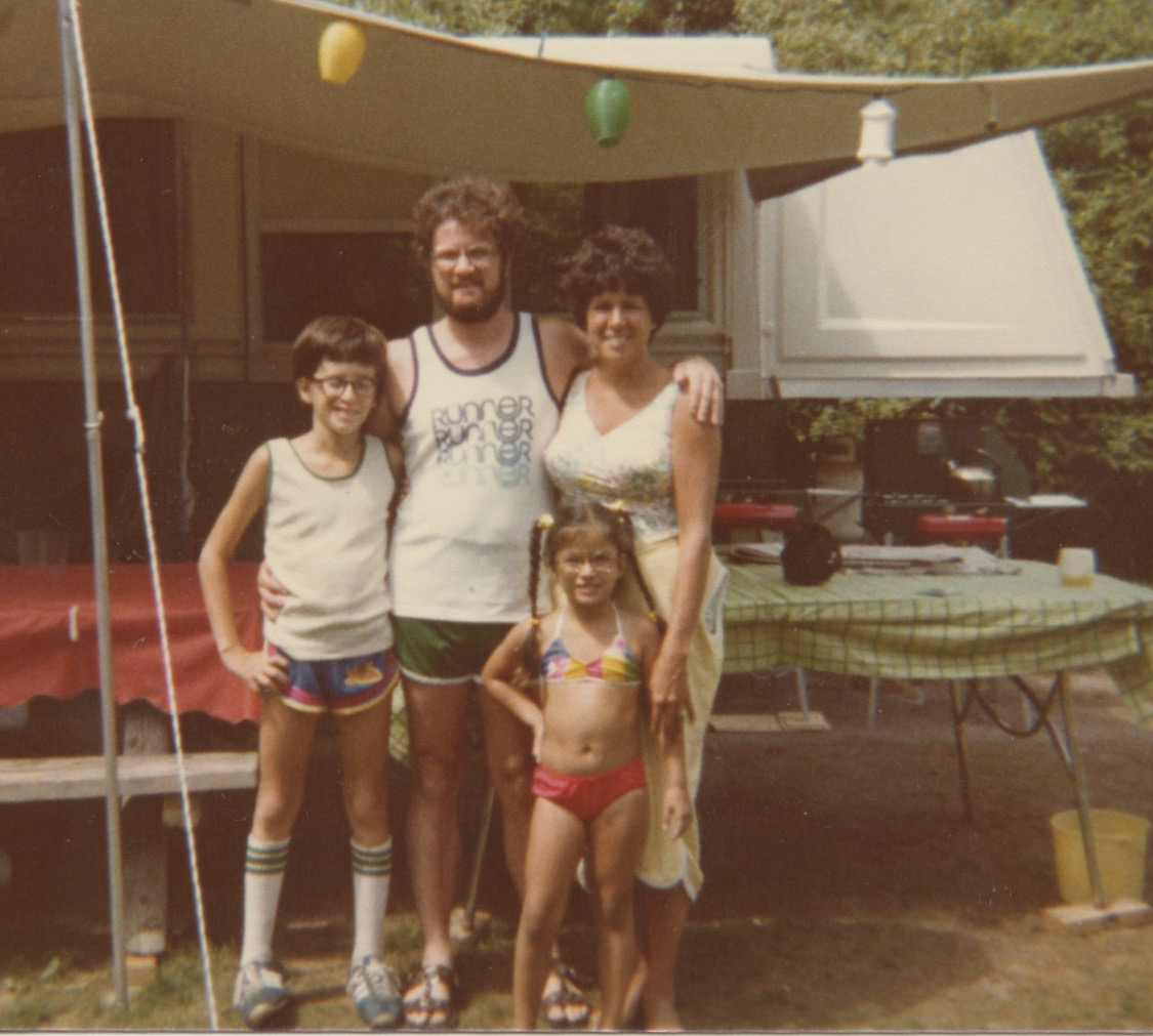 A young white family stand in front of tables set up in front of their pop-up camper. The young boy is wearing tube socks and sneakers with blue running shorts and a tank top. The older man is also wearing a tank top with the word runner repeated down the front of it. A little girl with pigtails stands in front in a bikini. Her mother, a white woman with a tan, stands behind her.  