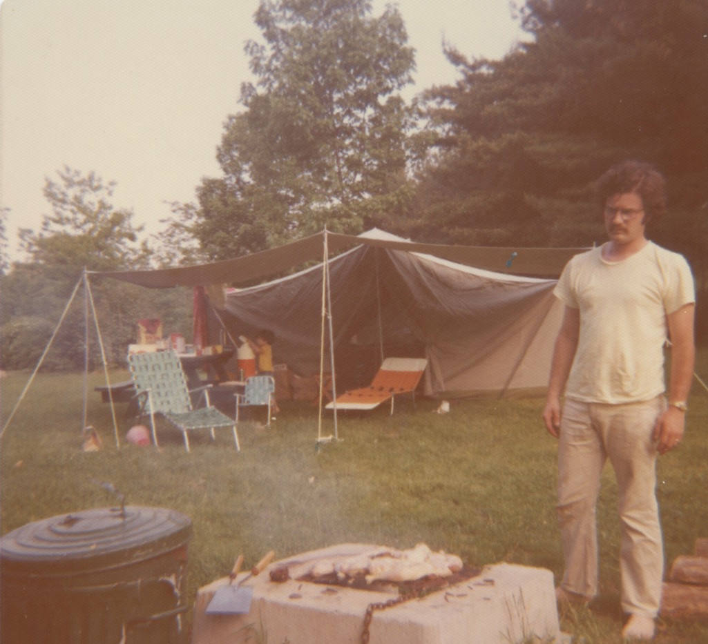 A large tent is set up in the background. In front of it are two outdoor chaises and one child-size lawn chair in front of a picnic table. In the foreground. a young man in a t-shirt and long pants stares at a lit grill with chicken pieces cooking on top of it.