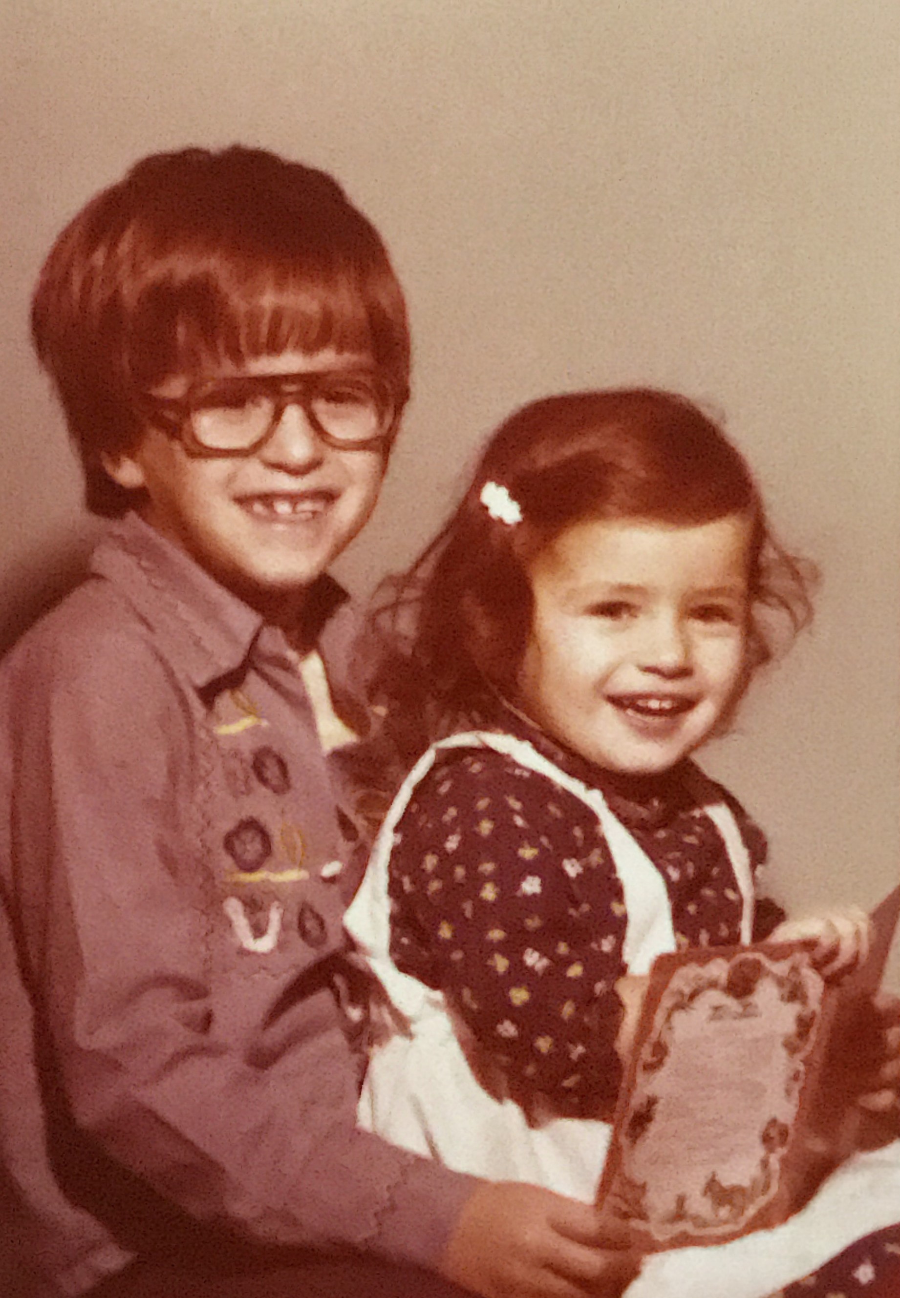 Two young white children. The boy is taller with dark hair in a bowl cut, wears glasses, and has a gap-toothed smile. The girl has long dark haired pulled back to one side with a white barrette. She's wearing a white jumper with dark blouse underneath. Both children are holding a book that sits in the little girl's lap.
