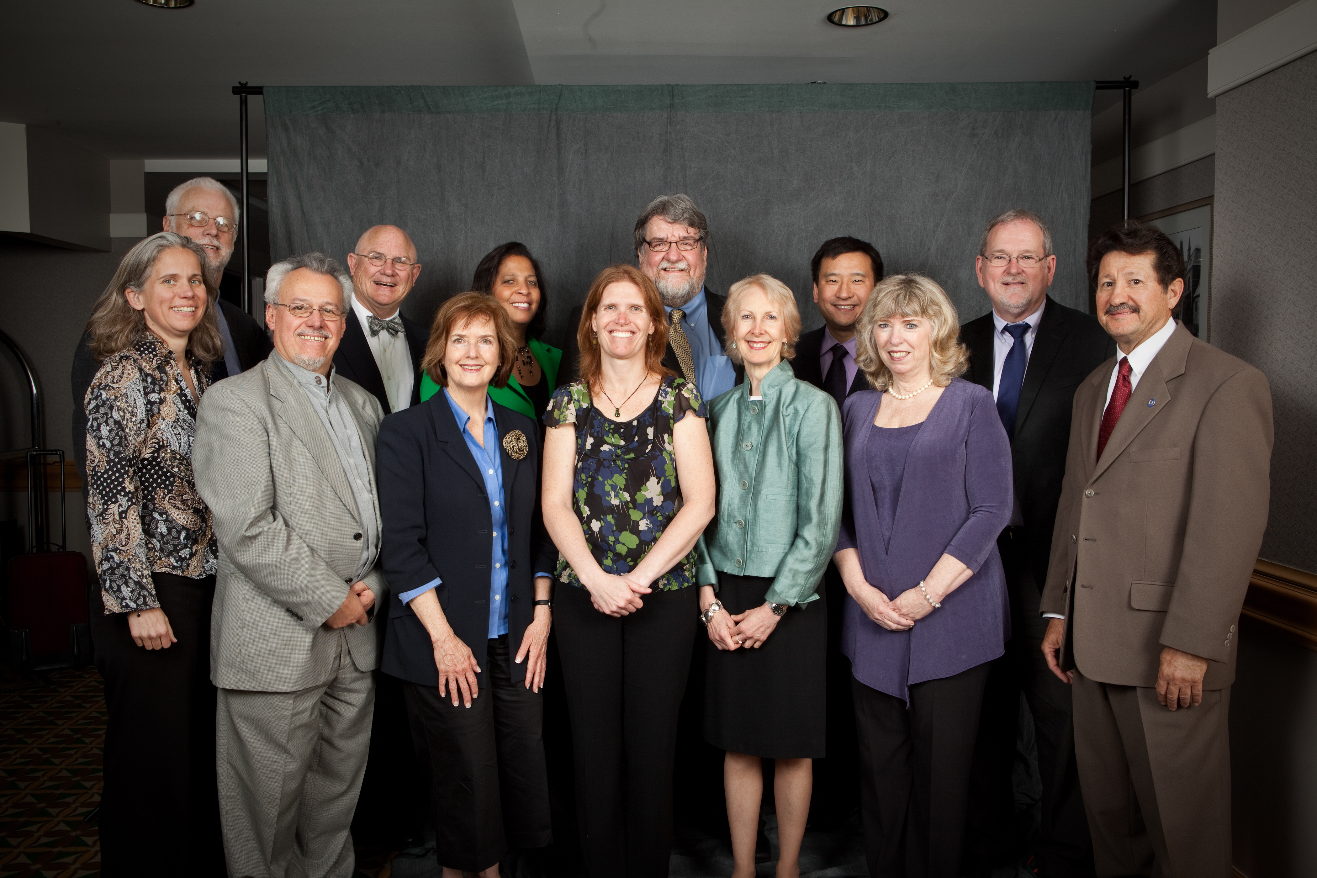 A group of middle-aged to older adults standing in a room. There are five white women,  one Black woman, one Asian man, and six white men. They are all dressed in business attire. 