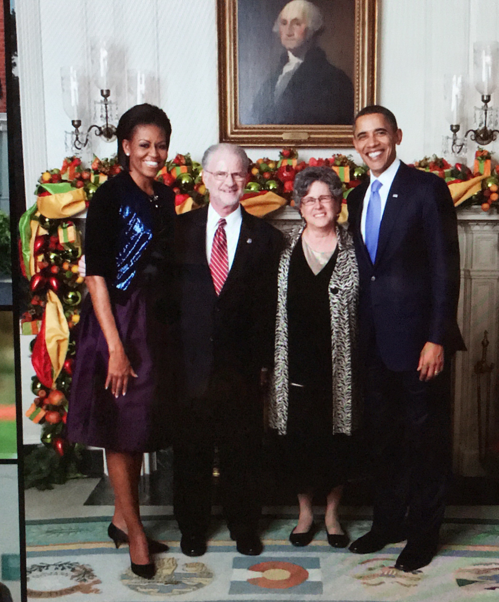 A tall, smiling, middle-aged Black woman stands next to a shorter older white man in a dark suit. His wife, an older white woman with wavy gray hair in a black dress with a gold and black jacket, stands next to him. On her other side is a tall middle-aged Black man in a dark suit with a light blue tie. The group is standing in front of a mantel decorated with ornaments and ribbon with a portrait of George Washington hanging above.