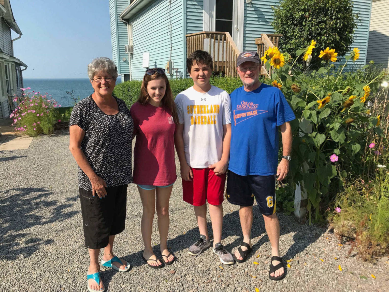 An older white woman with gray hair and glasses stands next to a young white girl with long brown hair and sunglasses on her heads. Next to her is a young white boy in a white t-shirt and red shorts and an older white man in a blue t-shirt with a baseball cap. Behind him is a planting of sunflowers in front of a blue house. In the distance you can see a lake.