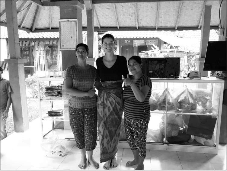 Erin, a tall smiling wadd alhite woman with dark hair wearing a patterned sarong, stands in a shelter containing display cases alongside two shorter Balinese women in striped t-shirts and patterned trousers.