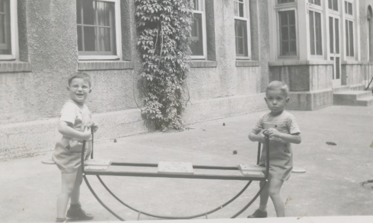 A black-and-white photo of two young white boys in a courtyard of a large concrete building. They are both sitting on a round seesaw. Both are facing the camera.Image