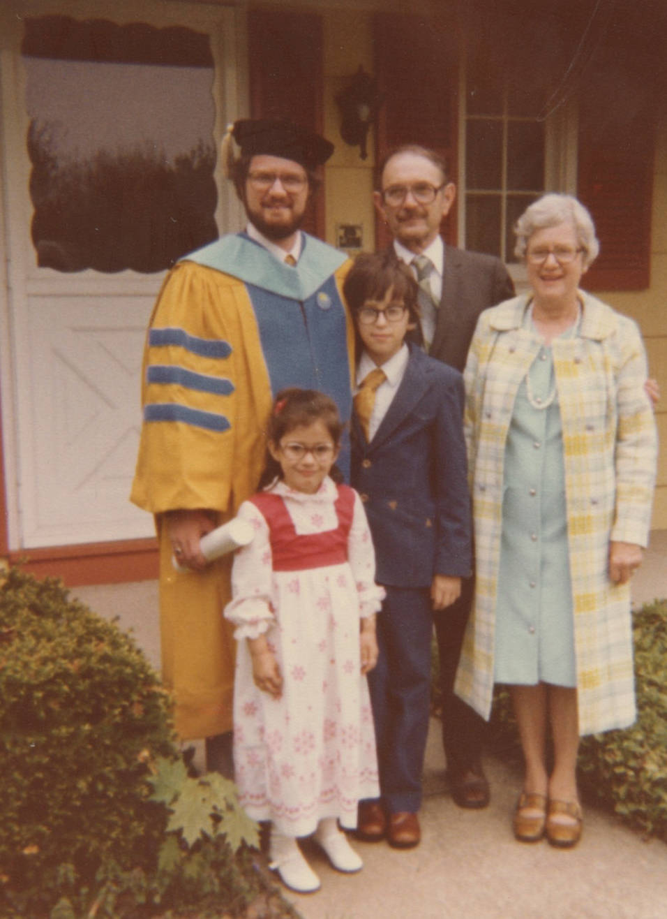A middle-aged white man in glasses wears a yellow robe with three blue stripes on the sleeve and a mortar board with a tassel on his head. He's holding a white tube in one hand. Next to him are an elderly white man in a gray suit and an older white woman with white hair wearing a light blue dress with a blue and yellow coat. A young boy with glasses stands in front of the man in the robe. He's wearing a blue suit and yellow tie. Next to him is a little girl with glasses in a white and red long dress. 