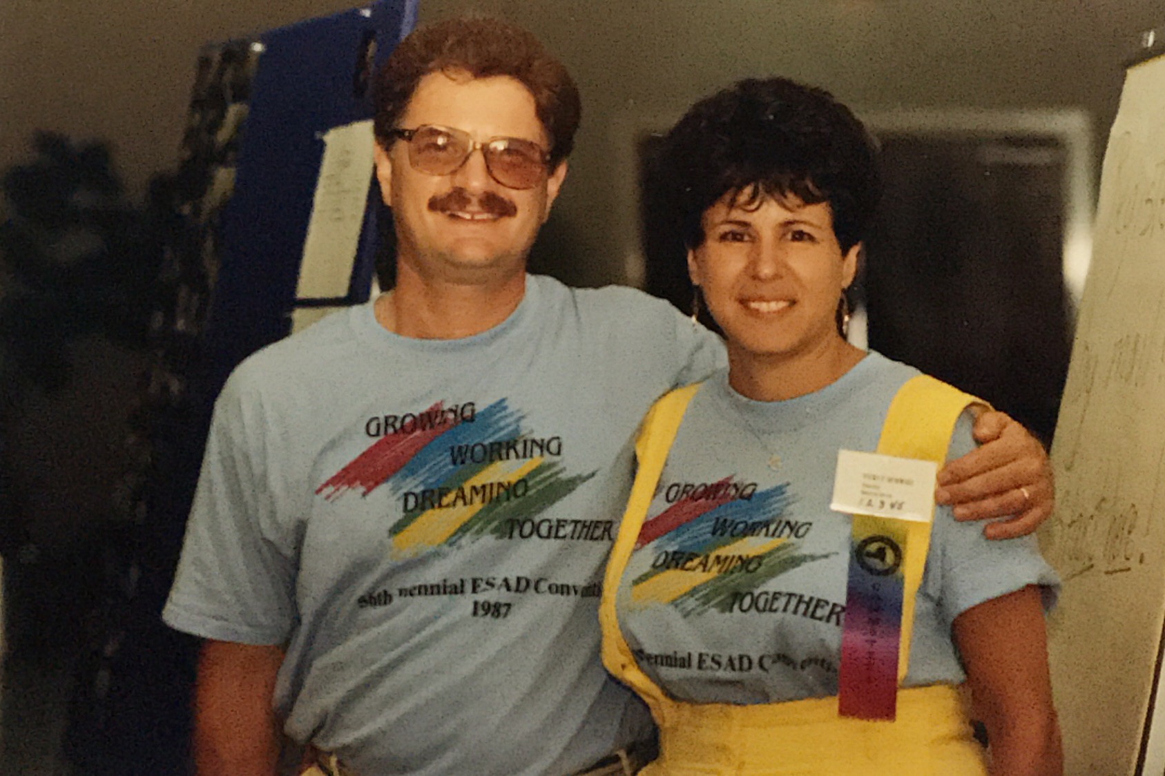 A young white man with short curly hair and tinted glasses wears a light-blue t-shirt with the words Growing Working Dreaming Together stands with his arm around a young white woman wearing the same t-shirt under a yellow jumper. She has dark wavy hair and is also wearing a name tag with a ribbon.