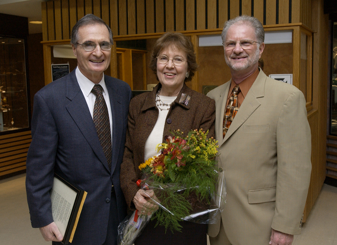 An older white man with dark hair and glasses wearing a dark navy suit and brown tie holds a plaque by his side. Standing next to him is a middle-aged white woman with reddish hair and glasses. She's holding a bouquet of yellow and red flowers. To her left is another older white man with white hair and a white beard in a tan suit with an orange button-up shirt. All are smiling for the camera.