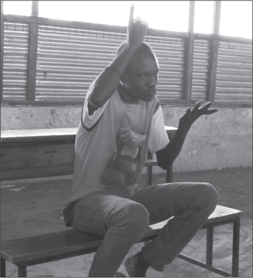 Sitting on a wooden bench in a wood and concrete building, Atem, a young black man in jeans and a faded T-shirt, signs VILLAGE (right fist and raised index finger on his head).