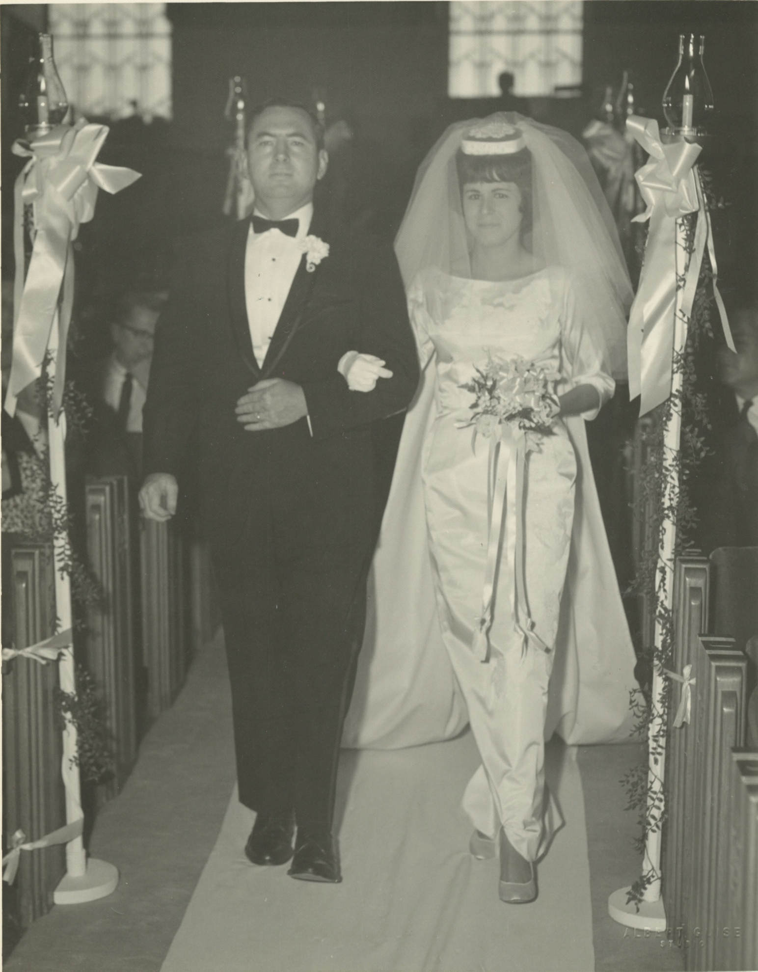 A middle-aged white man in a dark colored tuxedo escorts a young white woman down the aisle at a wedding. She is wearing a long white dress with a train and a veil on her heard. She's hold a bouquet of flowers with long dangling ribbons. She is holding on to the middle-aged man's arm.