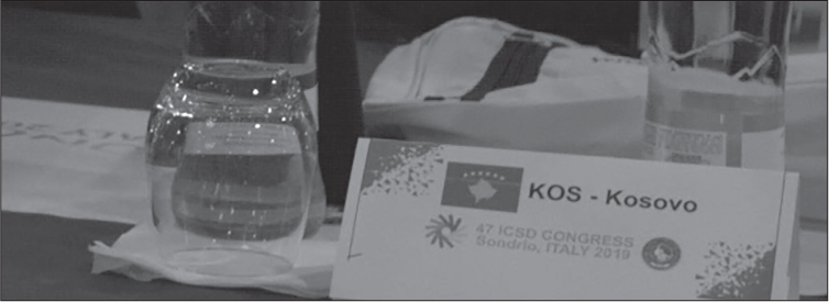 Close-up of a trestle table with a glass of water, various papers, and a place marker reading KOS – KOSOVO 47 ICSD CONGRESS, Sondrio, ITALY 2019.