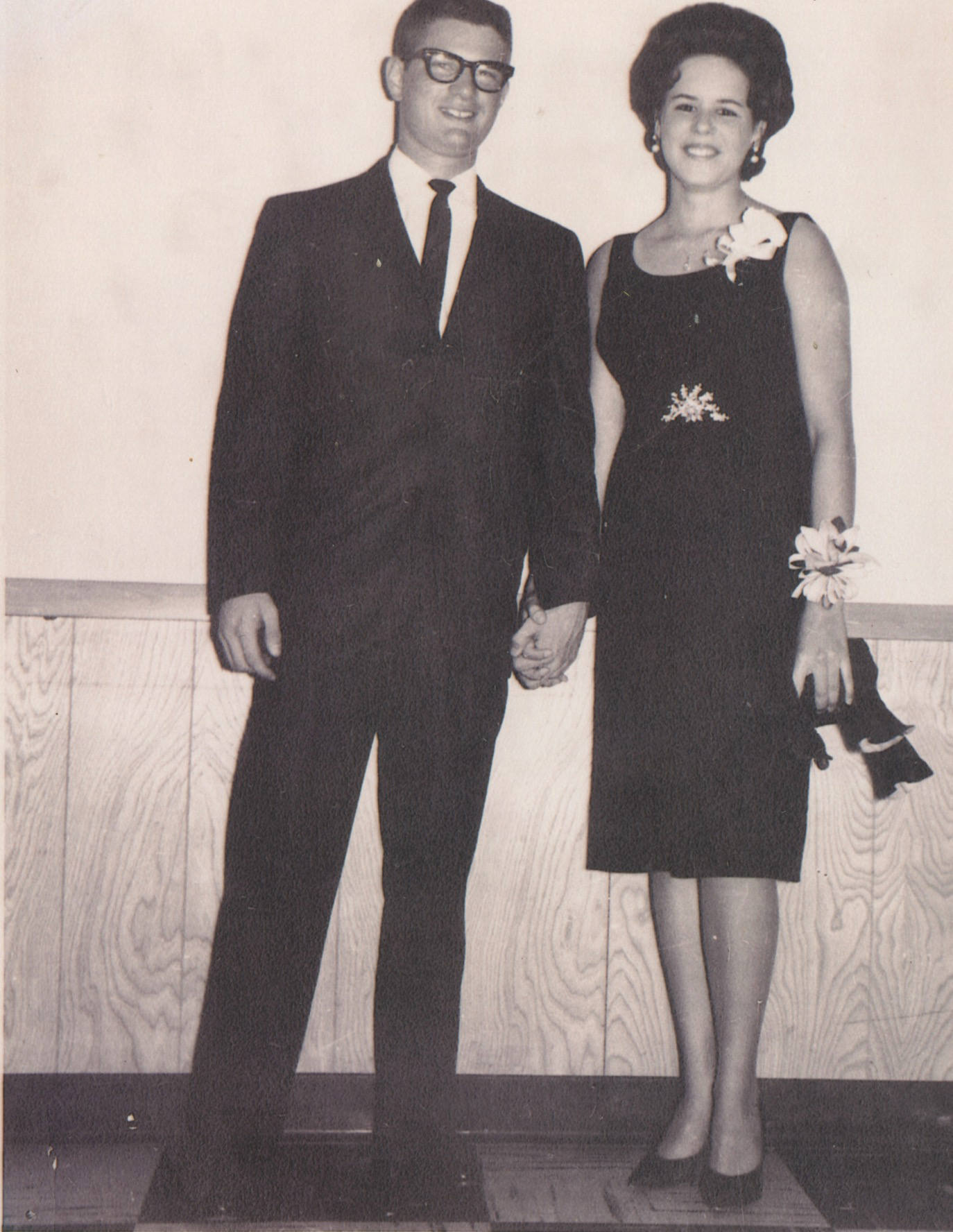 A young white man and a young white woman holding hands. He's wearing a dark suit and tie and has horn-rimmed glasses. She's in a dark colored sleeveless dress and has a wrist corsage.