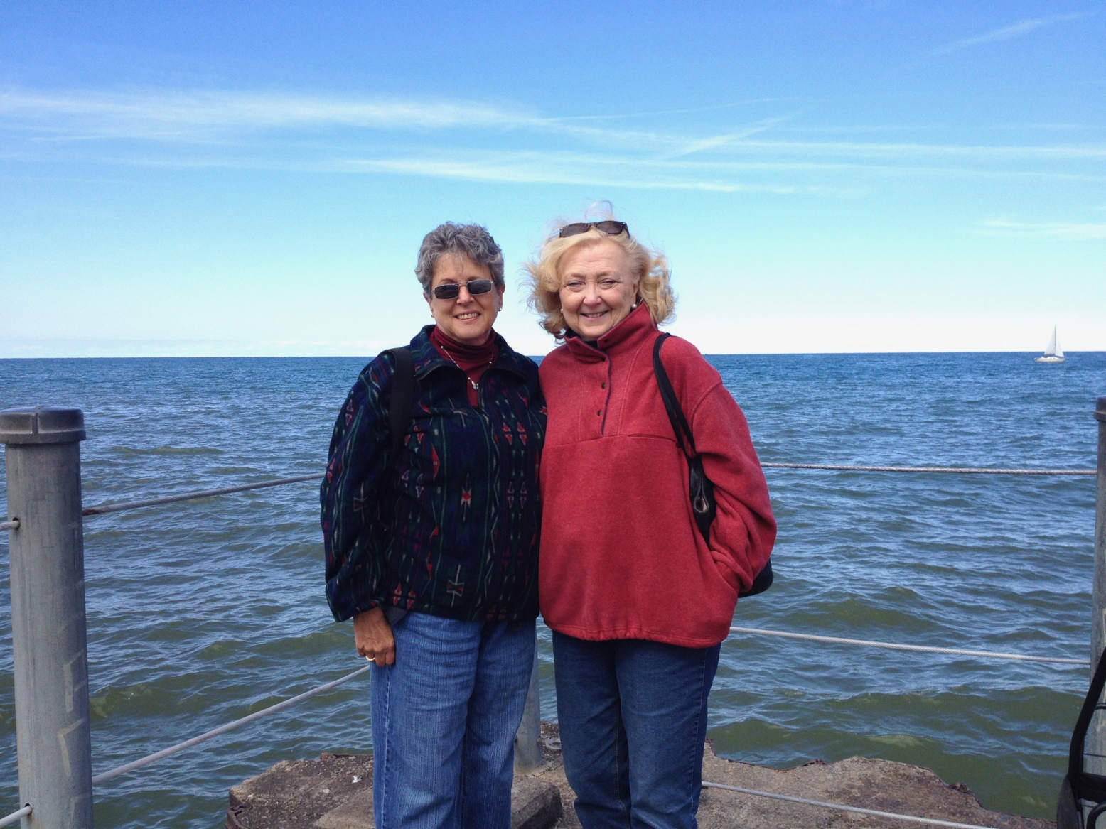 Two older white women standing on a pier on Lake Ontario. Far in the distance, a sailboat crosses the lake. The woman on the left has short wavy gray hair and is wearing sunglasses, a dark top, and jeans. The woman on the right has on a red top and jeans. Her sunglasses are on top of her head and she's carrying a purse. 