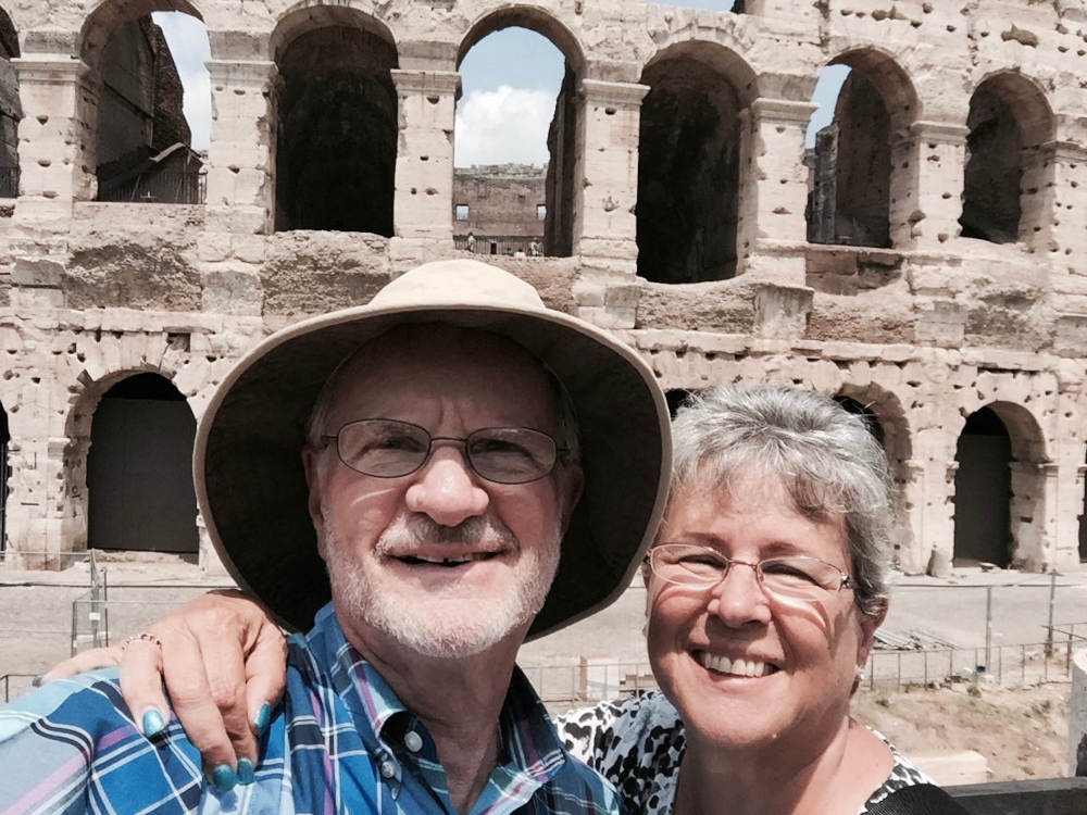 An older white man and woman stand in front of the stone coliseum in Rome. He's wearing a hat, glasses, and a blue plaid shirt. She has short wavy gray hair and glasses and is wearing blue fingernail polish.